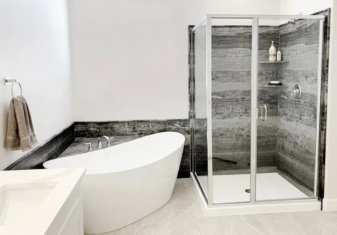 A newly remodeled residential bathroom with a new luxurious white bathtub and walk-in shower with glass doors, a stone surround that matches the tub area, storage shelves, and new modern hardware.