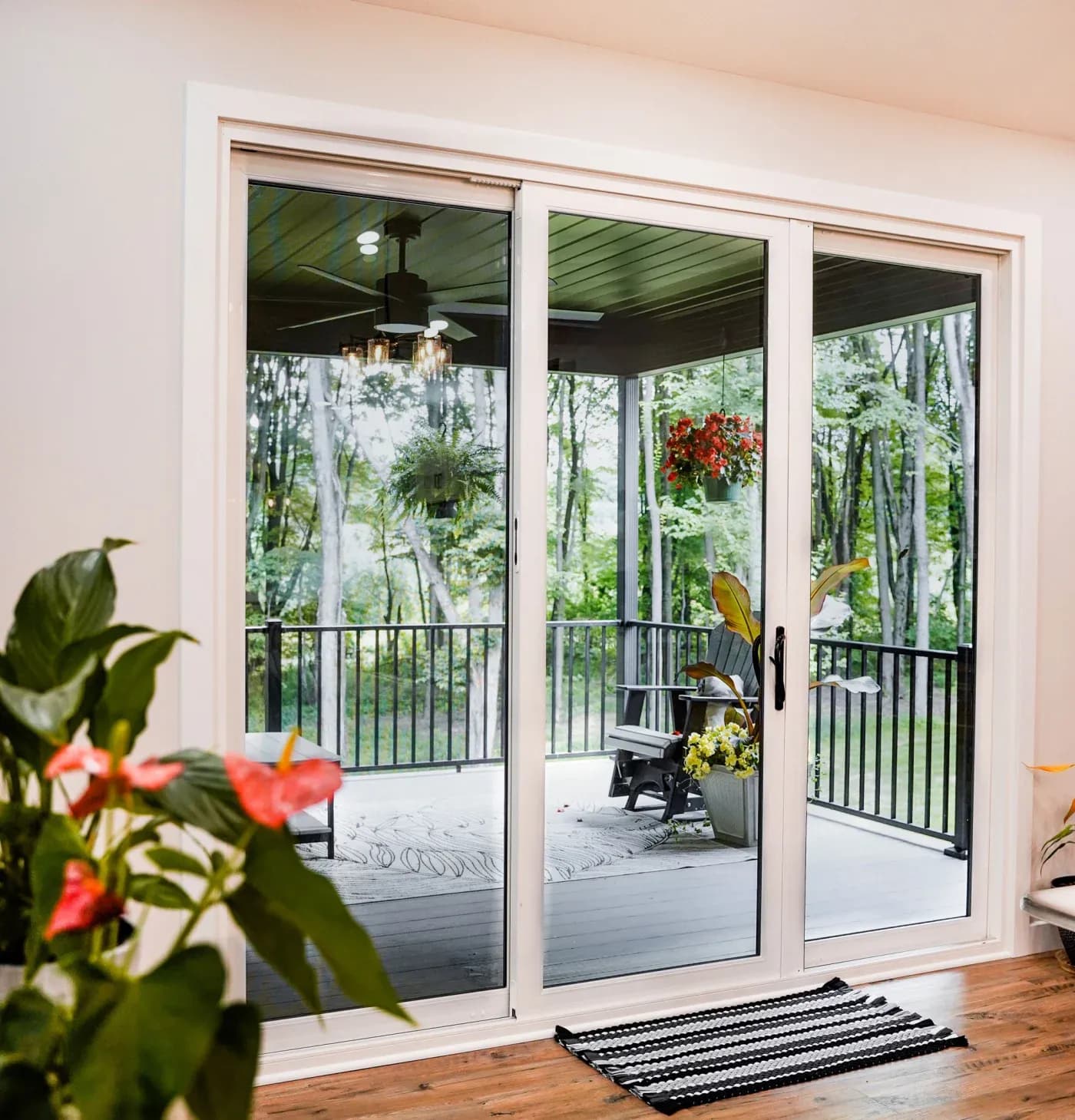 An inside view of a three-section white patio door overlooking a patio with metal railing, wood deck, lounging chairs, and planters with plants.