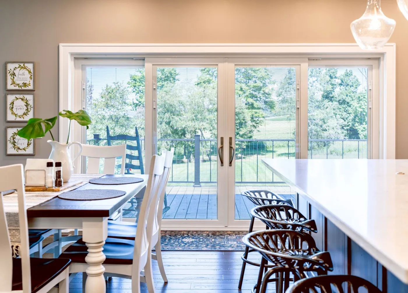 A dining room with a white dining table and chairs, a white kitchen counter and more chairs, furnishings, and a 4-lite patio door in white with internal blinds.