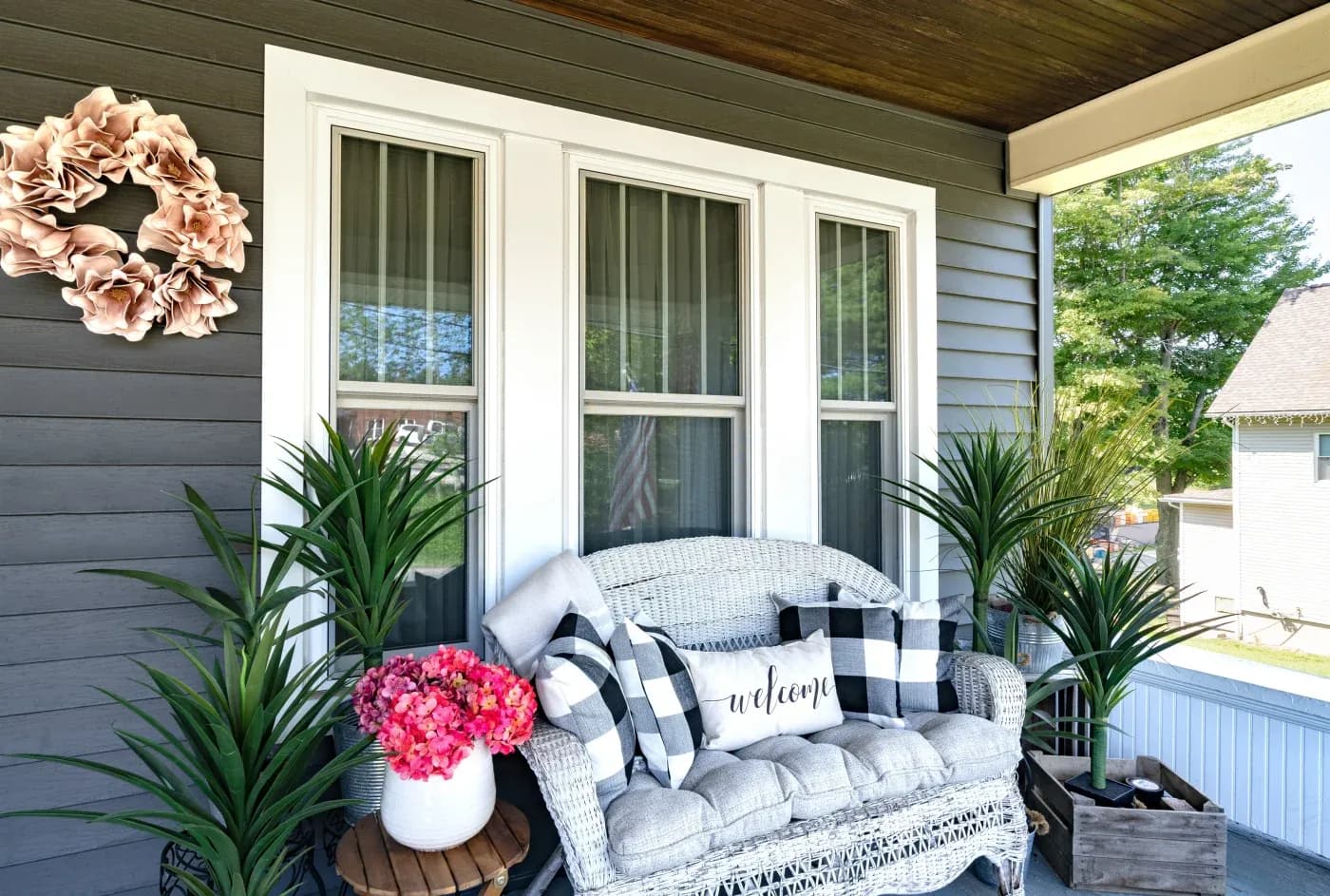 A section of a residential front porch in NW Arkansas with lots of potted plants around a small seating area with a row of three white double-hung windows in the background against the brown panel siding.