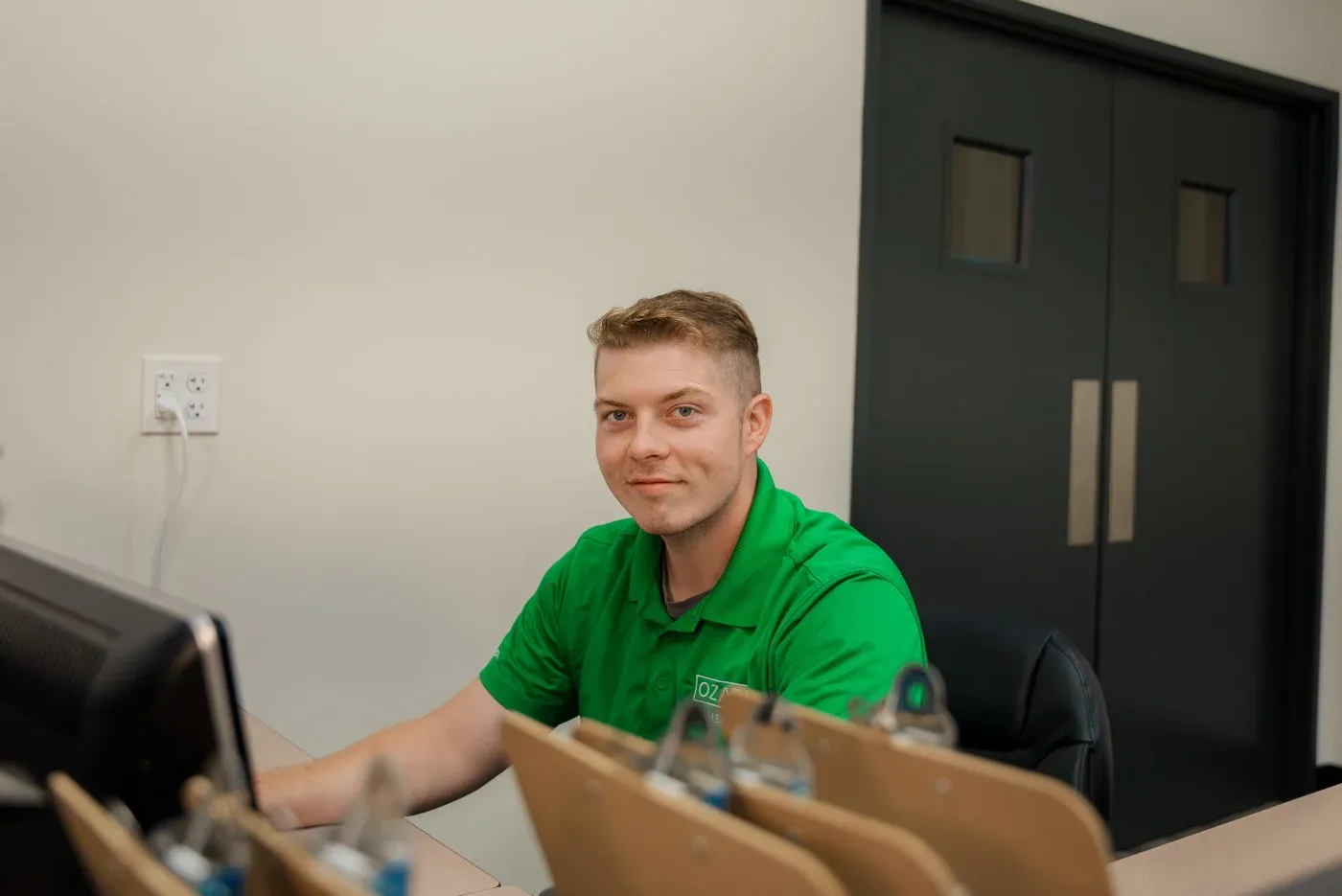 An Ozark Home Pros production manager sitting at his desk in their Springdale, AR design center looking at the camera in his company shirt and smiling.