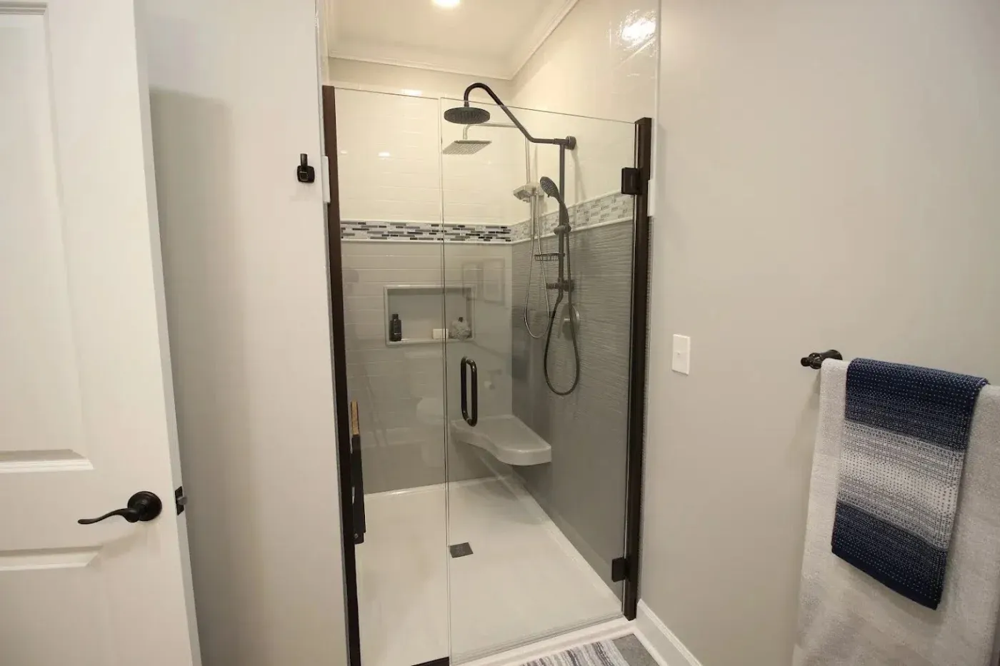 A new residential walk-in shower with glass doors and black trim, seating, storage shelving in the back wall, and new modern hardware, including a handheld showerhead.