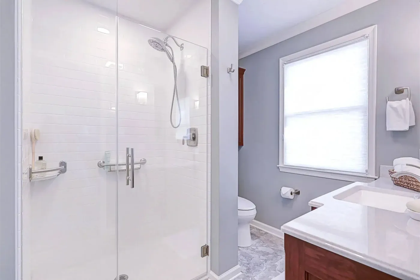 A large white and light blue residential bathroom with a new walk-in shower with white surround, two different grab bar shelves, glass doors, and new modern hardware, including a hand-held showerhead.
