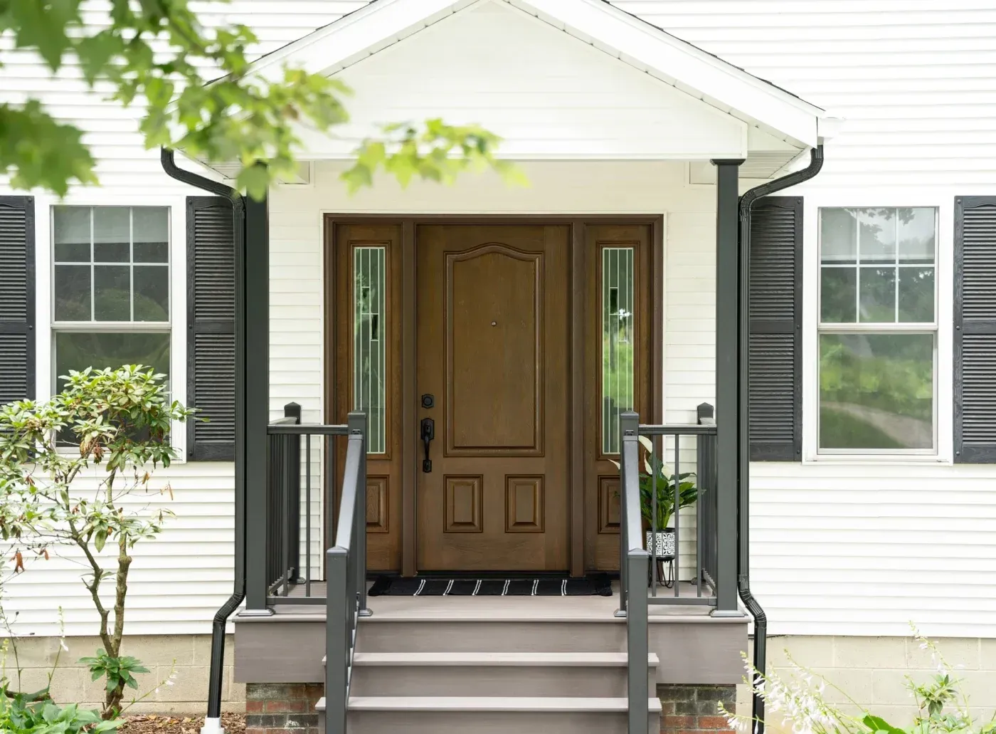 A close-up shot of the front of a home with white panel siding, dark green trim and gutters, and one double-hung window on each side of the entryway with a new entry door with natural bark style and a Cherry finish.
