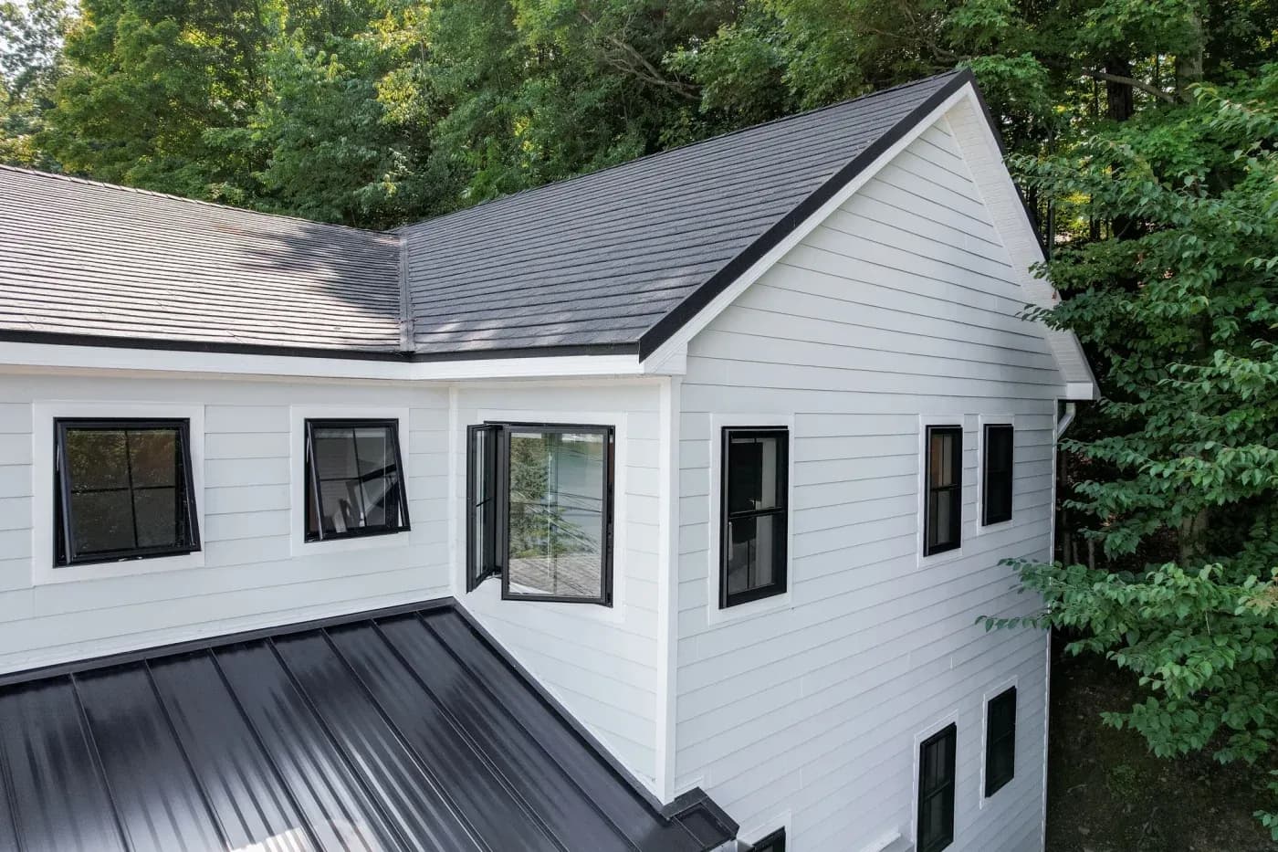 Outside of a large home with many replacement windows, including awning with textured matte coal black trim in contrast to the white plank siding and black metal roofing.