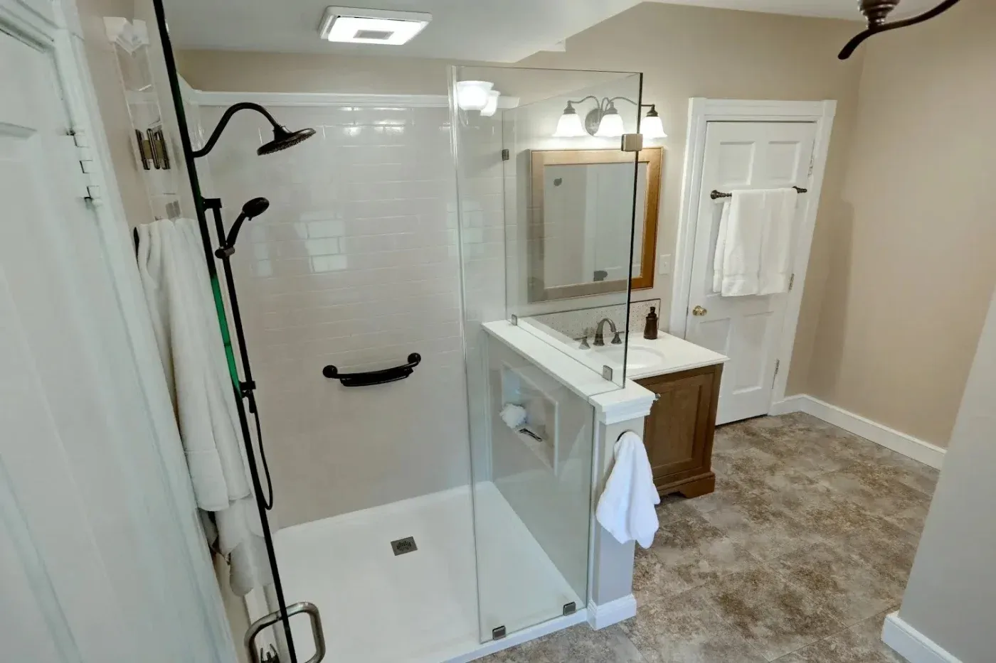 A large residential bathroom in NW arkansas with a new walk-in shower that’s white with black grab bars and hardware next to a small sink and bathroom doors, with stone tile flooring.