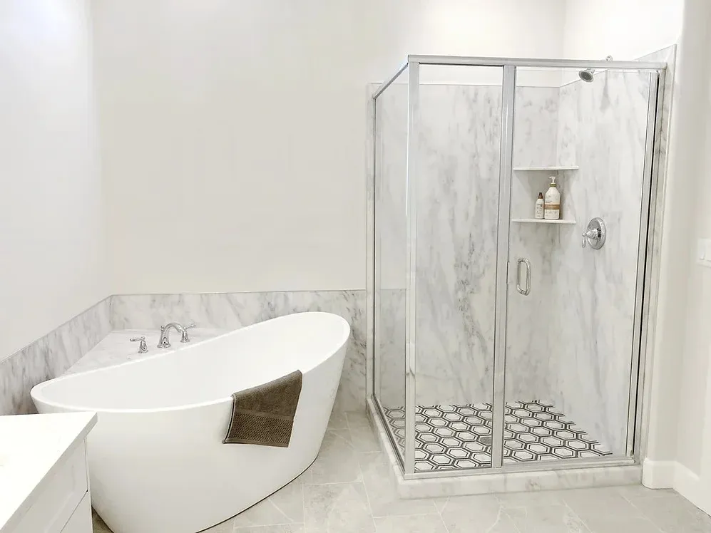 A small remodeled residential bathroom with white walls, a new luxurious-looking bathtub, and a walk-in shower next to it with a glass surround and unique tile flooring.
