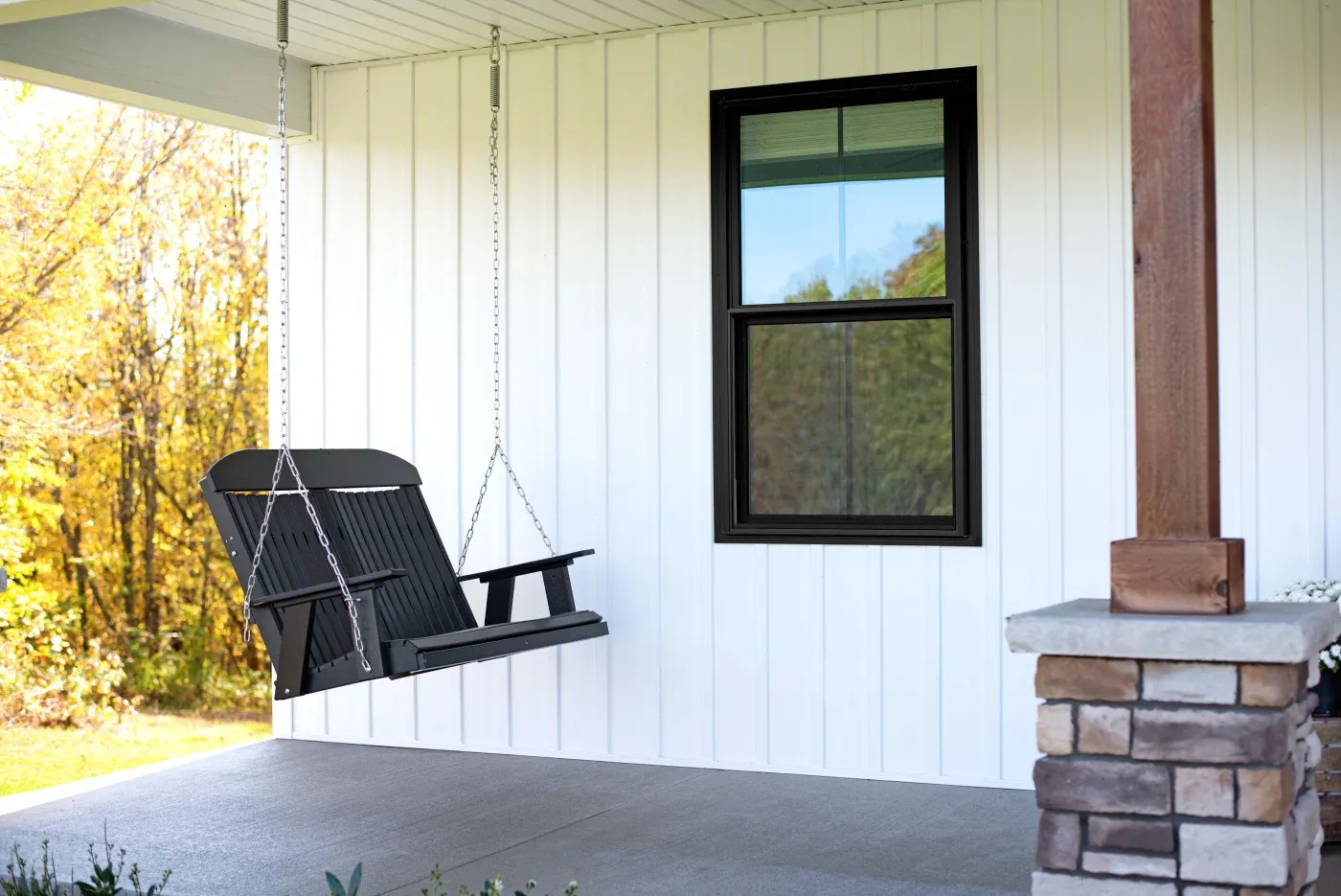 A front porch with a swinging chair, vertical white metal siding, a stone floor, and a double-hung window in classic bronze.