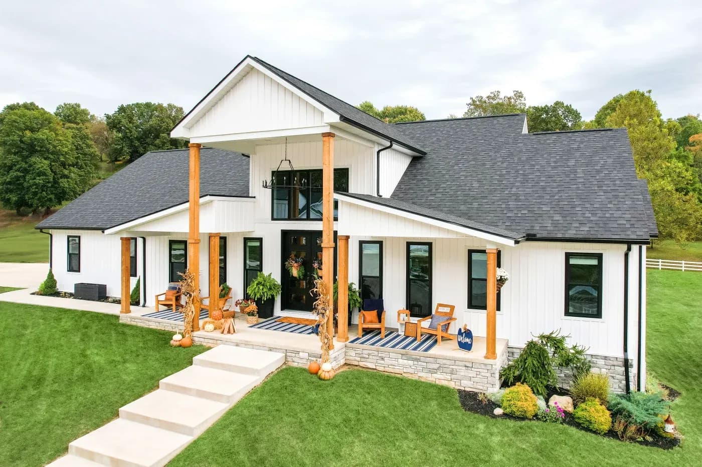 A beautiful home with a new roof, bright white vertical plank siding, black trim and gutters, and a variety of double-hung and picture windows. Bright green grass with trees in the background. Lounging chairs, planters, colorful white and blue rugs.